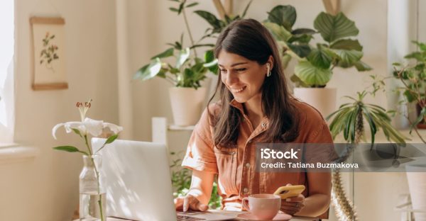 Positive young mixed race woman using a laptop and smartphone at home.Cozy home interior with indoor plants.Remote work, business,freelance,online shopping,e-learning,urban jungle concept.